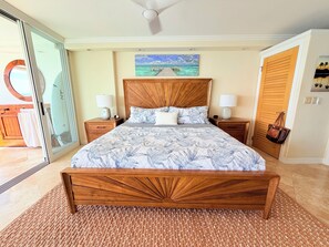 Un dormitorio con una cama de madera, dos mesitas de noche, una lámpara de noche, un ventilador de techo, una puerta corrediza de vidrio y un cuadro con una playa.