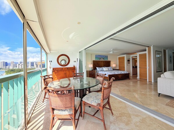 Un balcón con mesa y sillas, un dormitorio con cama y mesitas de noche, y vistas del horizonte urbano.