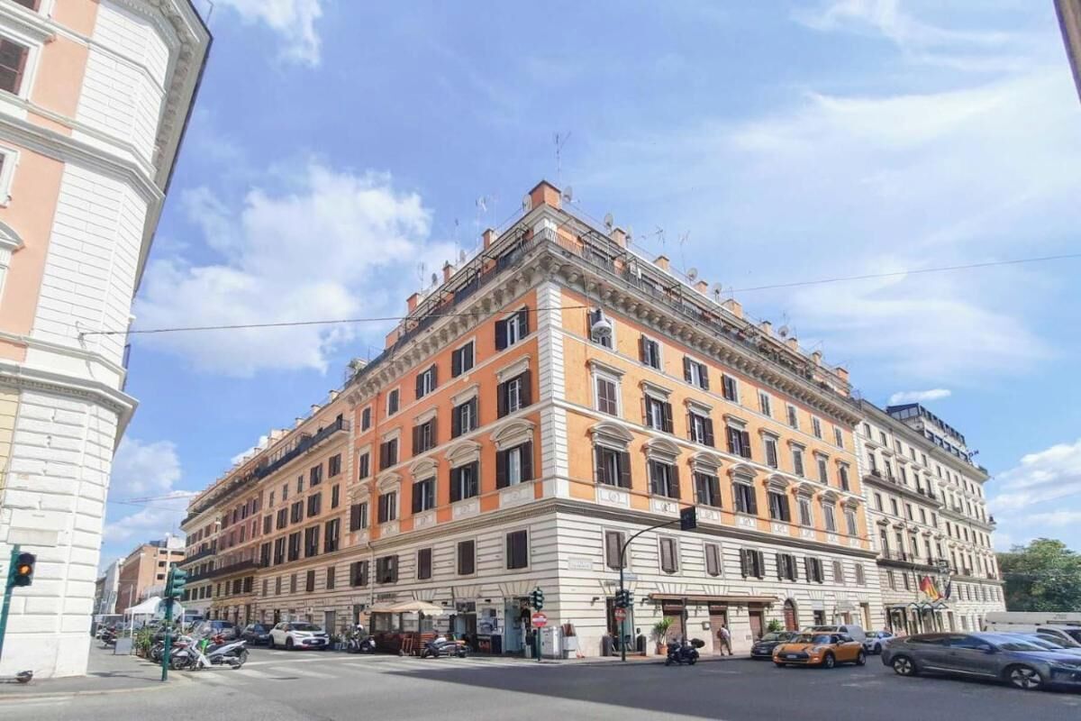 En byggnad med flera våningar, utformad i en klassisk arkitektonisk stil, med en central entré som har ett tak över, flera fönster och ett hörnläge med ett rundat hörn.