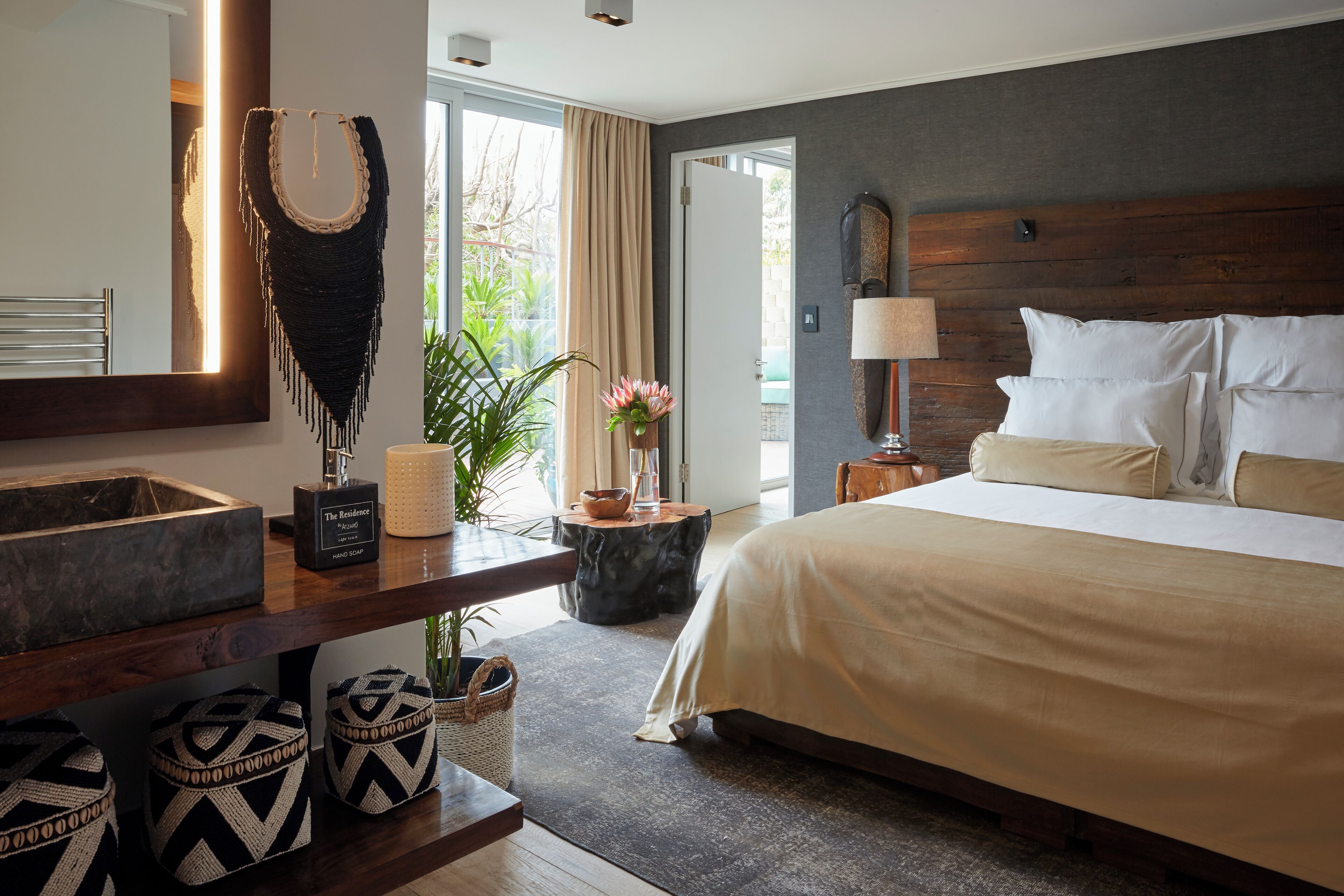 A modern hotel room with a large bed, a wooden headboard, a bedside table with a lamp, a large window with curtains, a bathroom with a mirror, and decorative pillows.