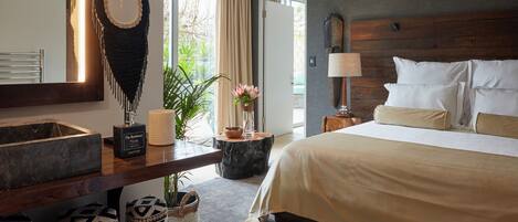 A modern hotel room with a large bed, a wooden headboard, a bedside table with a lamp, a large window with curtains, a bathroom with a mirror, and decorative pillows.