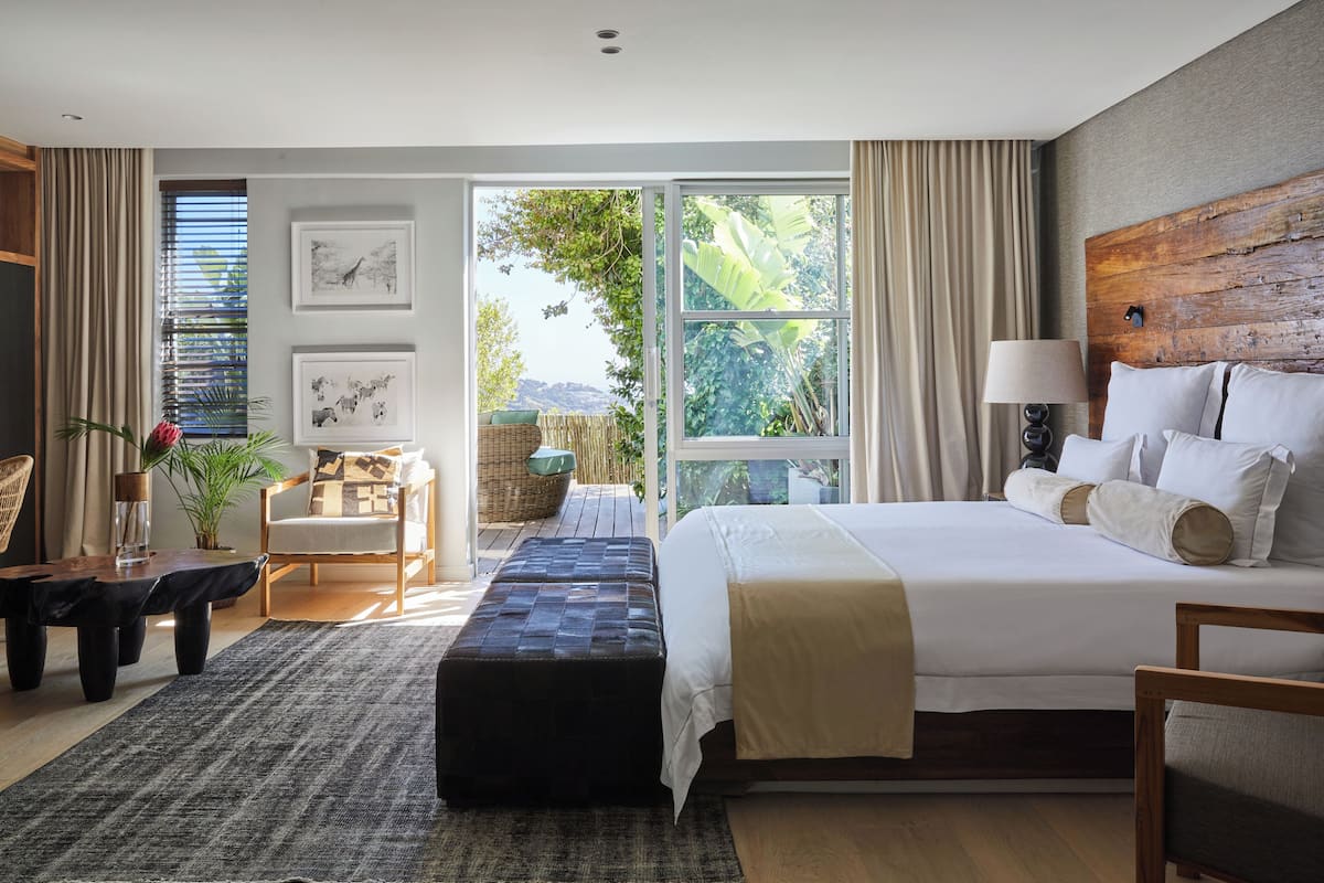 Ein geräumiges Schlafzimmer mit einem großen Bett, einem Holz-Kopfstück, Blick ins Grüne durch das Fenster, einer Sitzecke mit Stuhl und Tisch sowie einem kleinen Balkon.