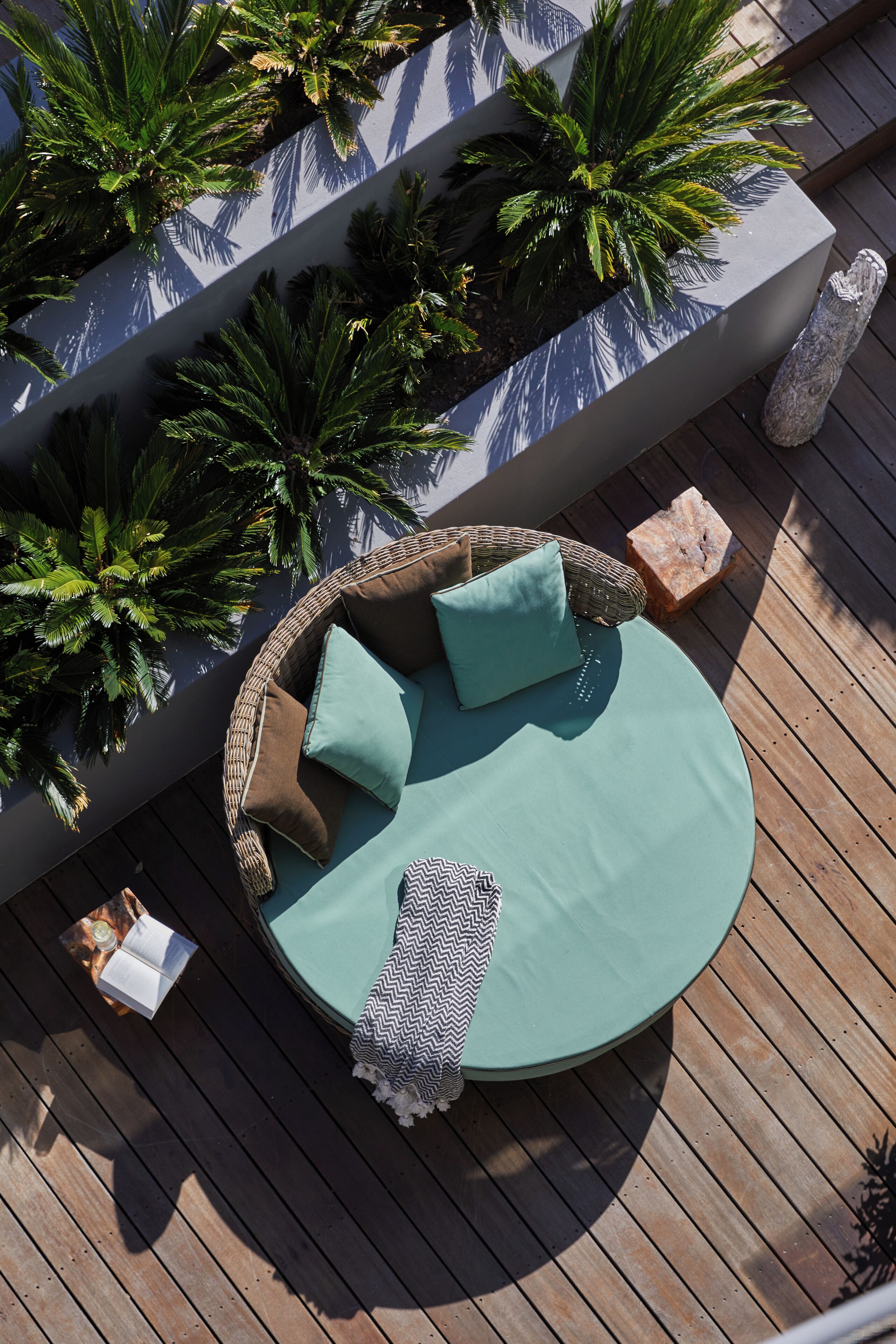 Un espacio de descanso al aire libre con cojines y un banco de mimbre, rodeado de macetas con plantas y una terraza de madera.