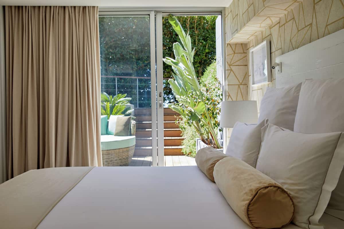 Ein Schlafzimmer mit Bett, einem Fenster mit Vorhängen und Blick auf einen Balkon mit Pflanzen und einem Stuhl.