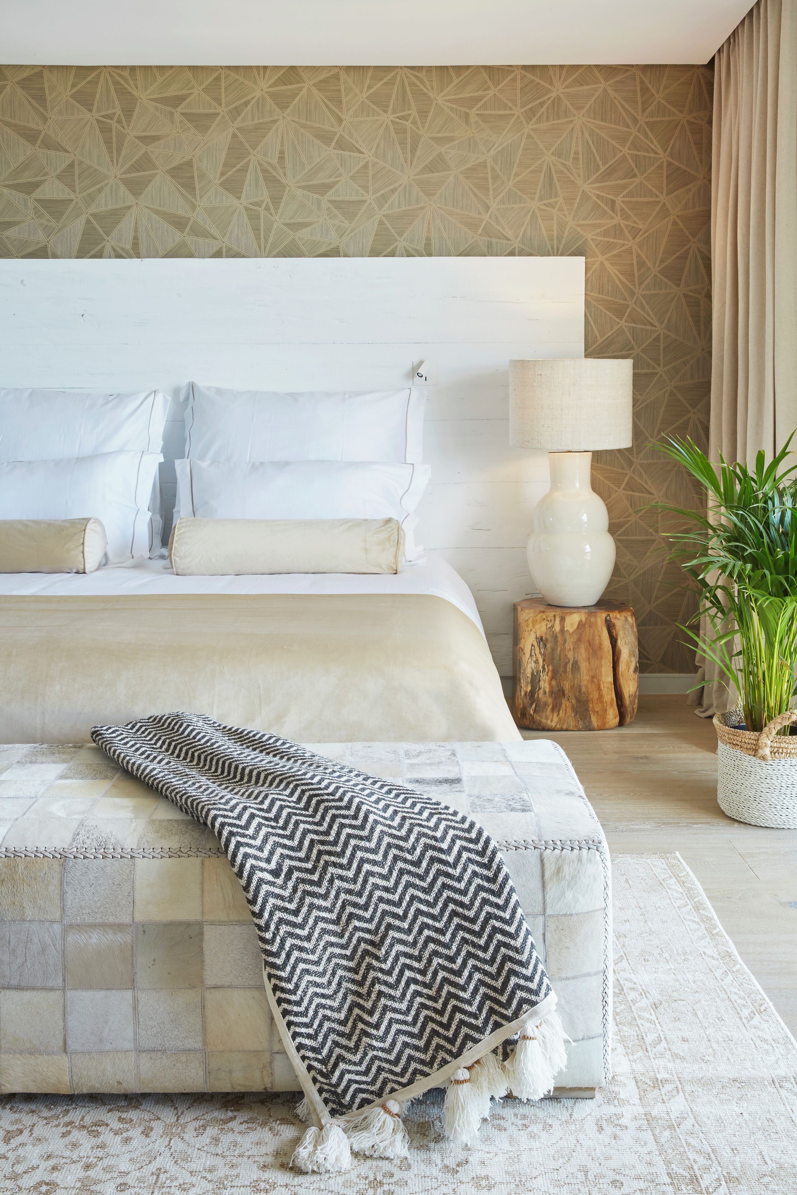 Un dormitorio moderno con una cama grande, una lámpara de noche, una planta en maceta y una pared con estampado geométrico.