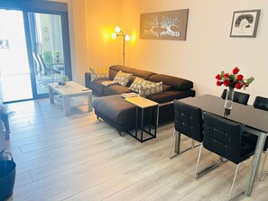 A modern living room with a sofa, coffee table, and dining table. There is a wall art piece and a vase with flowers. - Groundfloor Oasis Beach II La Zenia (Orihuela Costa)