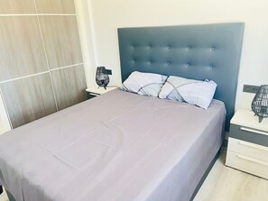 A neatly made bed with a grey bedspread and two pillows, a bedside table with a lamp, and a wall with a vertical wooden panel. - Groundfloor Oasis Beach II La Zenia (Orihuela Costa)