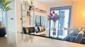 A modern living room with a glass dining table, a sofa, a TV, and a bookshelf with plants. - Groundfloor Oasis Beach II La Zenia (Orihuela Costa)