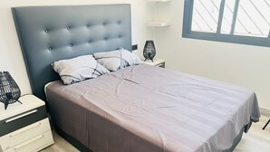 A modern bedroom with a tufted headboard, a bed with a striped pillow, a bedside table with a lamp, and a window with a view. - Groundfloor Oasis Beach II La Zenia (Orihuela Costa)