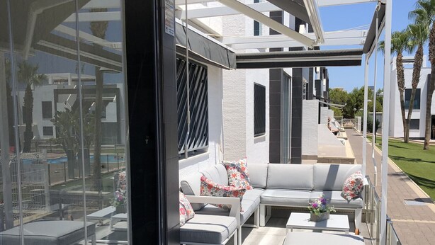 A modern outdoor patio with a glass enclosure, a grey sofa set, and a pool in the background. - Groundfloor Oasis Beach II La Zenia (Orihuela Costa)