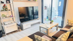 A modern living room with a large flat-screen TV, a sofa with patterned cushions, a coffee table, and a bookshelf with decorative items. - Groundfloor Oasis Beach II La Zenia (Orihuela Costa)