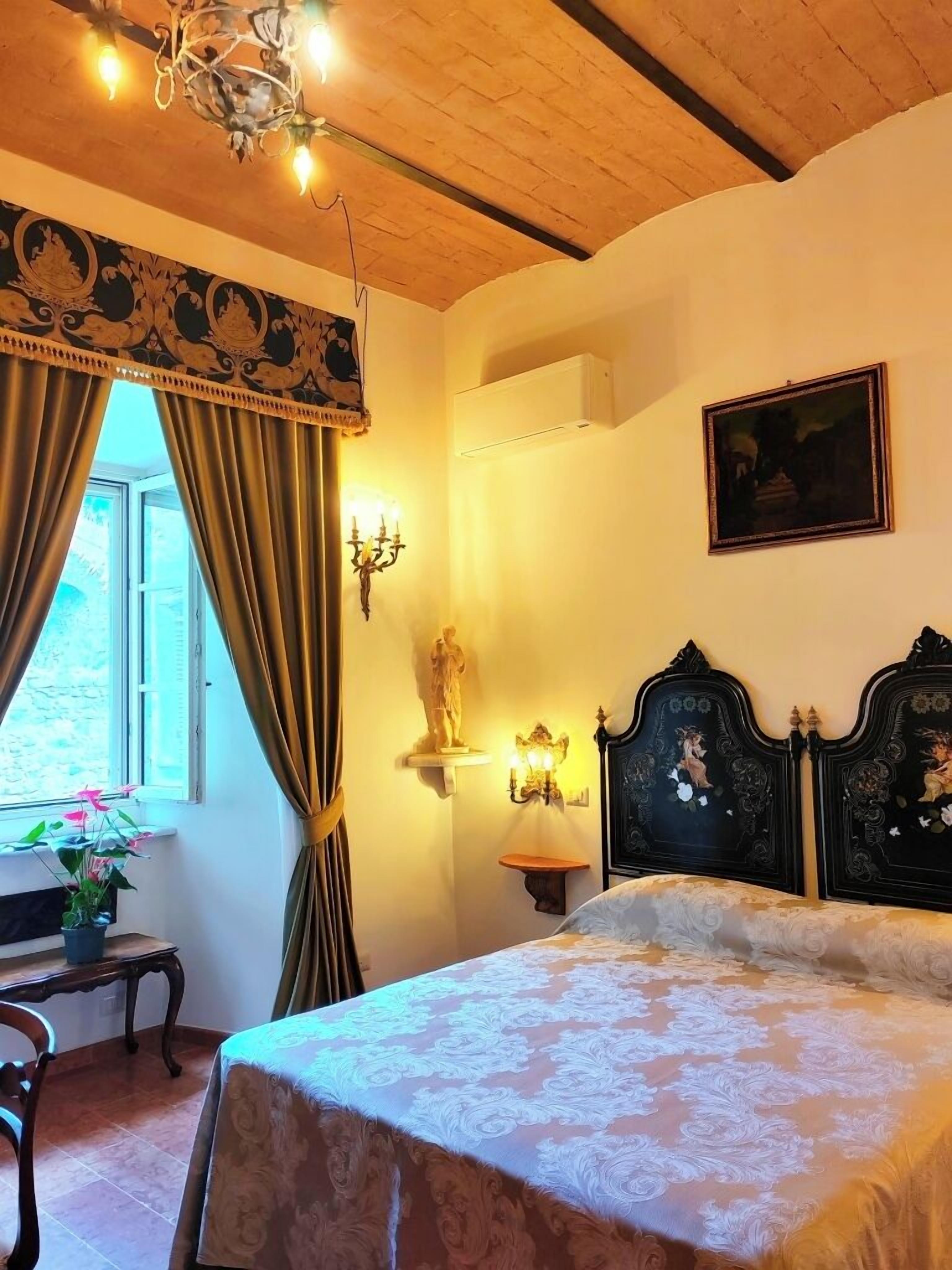 A bedroom with a bed, a chandelier, a window with curtains, a statue, and a painting on the wall.