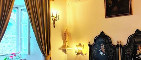 A bedroom with a bed, a chandelier, a window with curtains, a statue, and a painting on the wall.