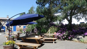 Outdoor wooden picnic tables with blue umbrellas, surrounded by potted plants and flowering bushes.