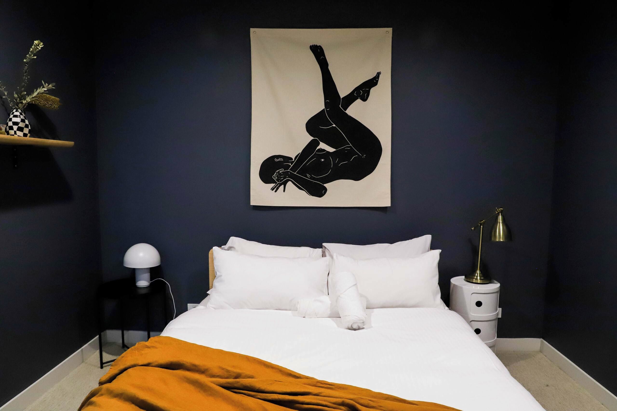 Une chambre à coucher avec un lit, des oreillers blancs, une couverture marron, une œuvre d’art représentant une personne, accrochée au mur, une table de chevet avec une lampe et une petite plante posée sur une étagère.
