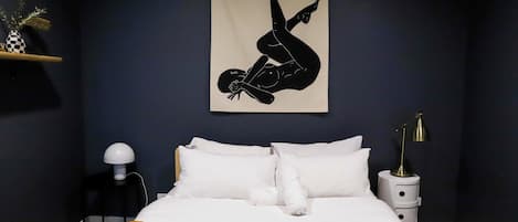 A bedroom with a bed, white pillows, a brown blanket, a wall-mounted artwork of a person, a bedside table with a lamp, and a small plant on a shelf.
