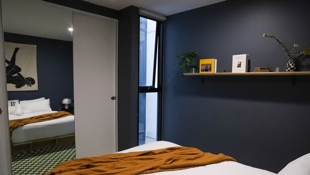 A hotel room with a bed, a bedside table, a mirror, a window, and a shelf with books and a plant. - Stylish 1BDR in the heart of Trendy Fitzroy (Melbourne)