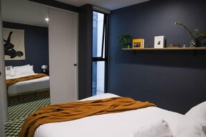 A hotel room with a checkered floor, a bed with a brown blanket, a white pillow, a shelf with books and a vase, and a window with a view.