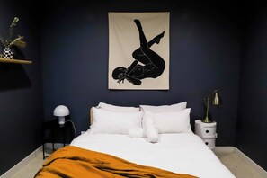 Une chambre à coucher avec un lit, des oreillers blancs, une couverture marron, une œuvre d’art représentant une personne, accrochée au mur, une table de chevet avec une lampe et une petite plante posée sur une étagère.