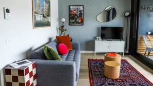 A modern living room with a grey sofa, colorful cushions, a mounted TV, and a large window with a view of buildings. - Stylish 1BDR in the heart of Trendy Fitzroy (Melbourne)