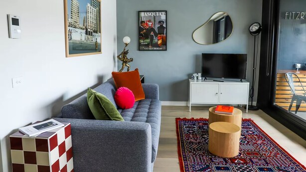 A modern living room with a grey sofa, colorful cushions, a mounted TV, and a large window with a view of buildings.