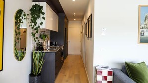 A modern kitchen with a central island, a dining area with a sofa and a green pillow, and a hallway leading to a door. - Stylish 1BDR in the heart of Trendy Fitzroy (Melbourne)