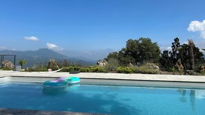 A swimming pool with inflatable rings, surrounded by a landscaped area with various plants and trees, and a view of distant mountains under a clear sky. - House with breathtaking views over the valley, only 15 minutes from Corte (Poggio-di-Venaco)