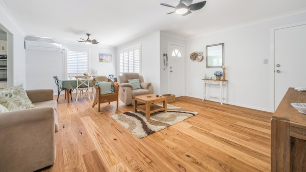 A spacious living area with a wooden floor, a beige sofa, a coffee table, and a dining table with chairs. There are ceiling fans, a white door, and a small side table with decorative items.
