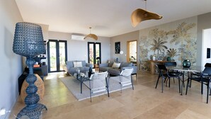 A spacious living area with a sofa, coffee table, and dining table, featuring a large window and a tropical wallpaper.
