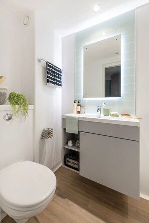 Un baño moderno con un inodoro blanco, un espejo sobre el lavabo y una pequeña repisa con artículos de aseo.