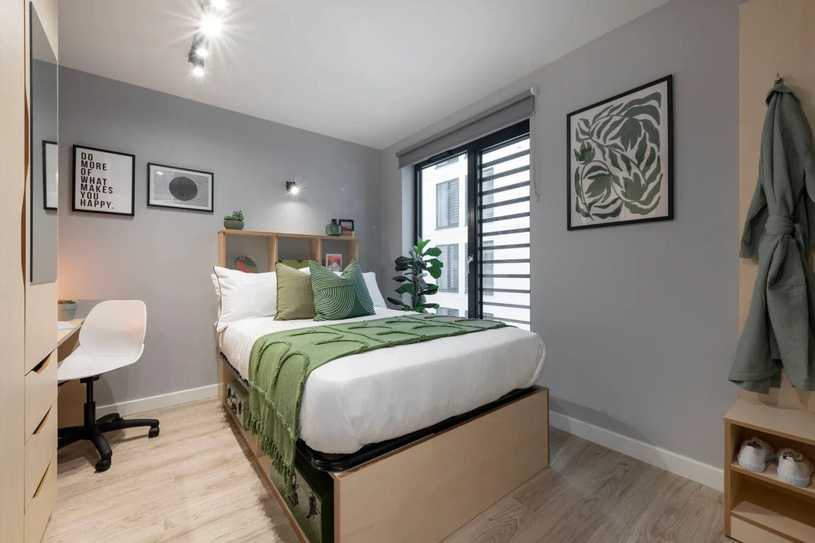 Un dormitorio moderno con cama, escritorio y silla. Hay dos cuadros en la pared, una ventana con persianas y una planta verde.