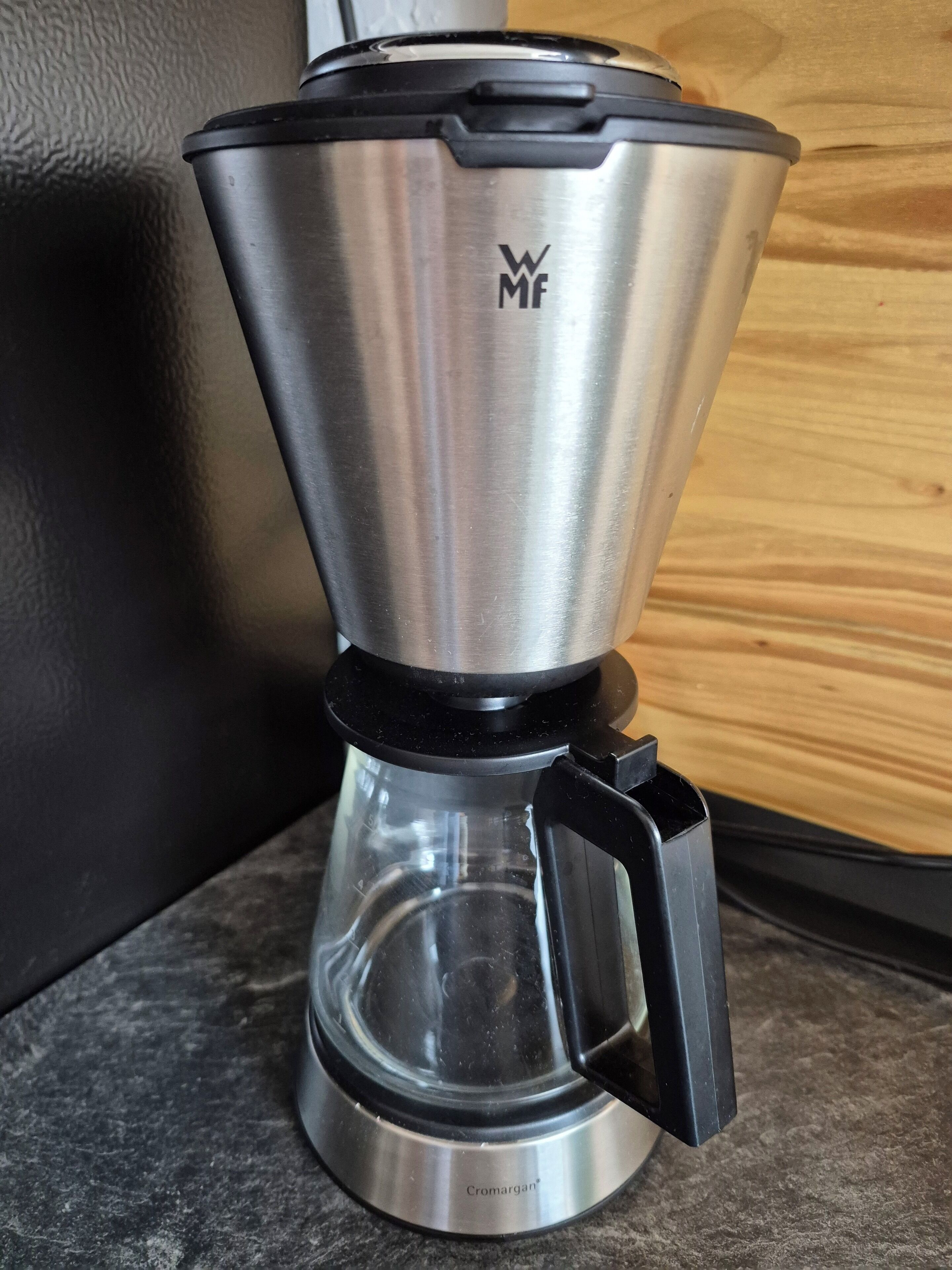 Une cafetière en acier inoxydable avec un manche noir et une verseuse en verre, posée sur un plan de travail.