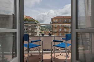 Un balcone con due sedie blu e un tavolino, affaccia su una zona residenziale con edifici e montagne sullo sfondo.