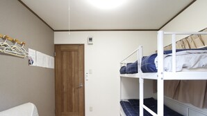 Een slaapzaal met stapelbedden, een houten deur, een kapstok met hangers en een prikbord met papieren.
