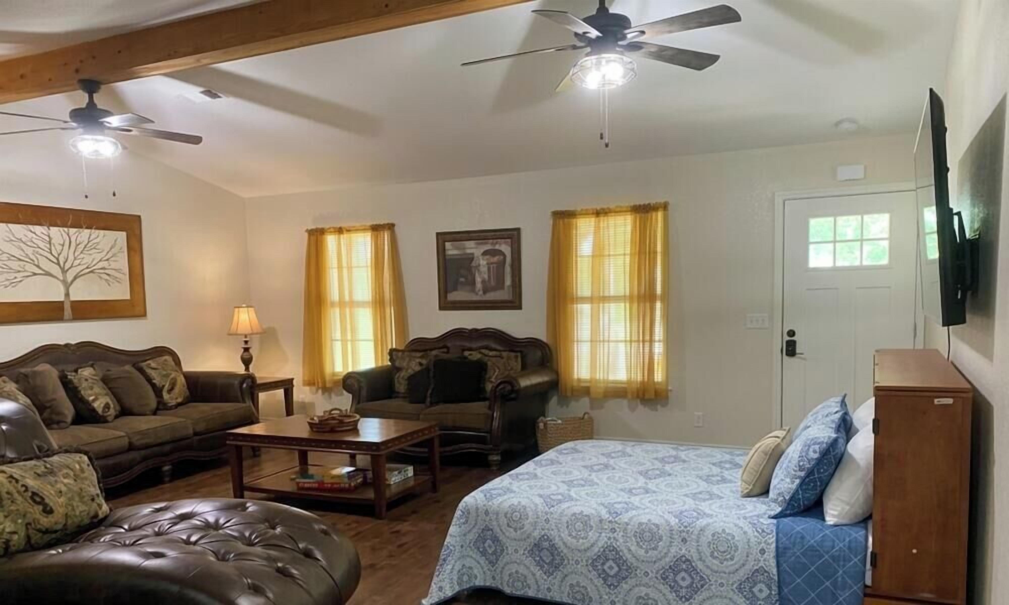 A living room with a sofa, a coffee table, a patterned bed, two windows with curtains, a ceiling fan, and a television mounted on the wall.