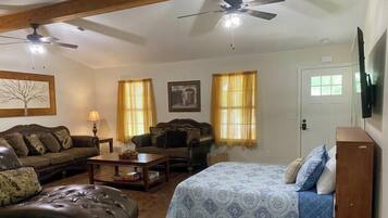 A living room with a sofa, a coffee table, a patterned bed, two windows with curtains, a ceiling fan, and a television mounted on the wall.