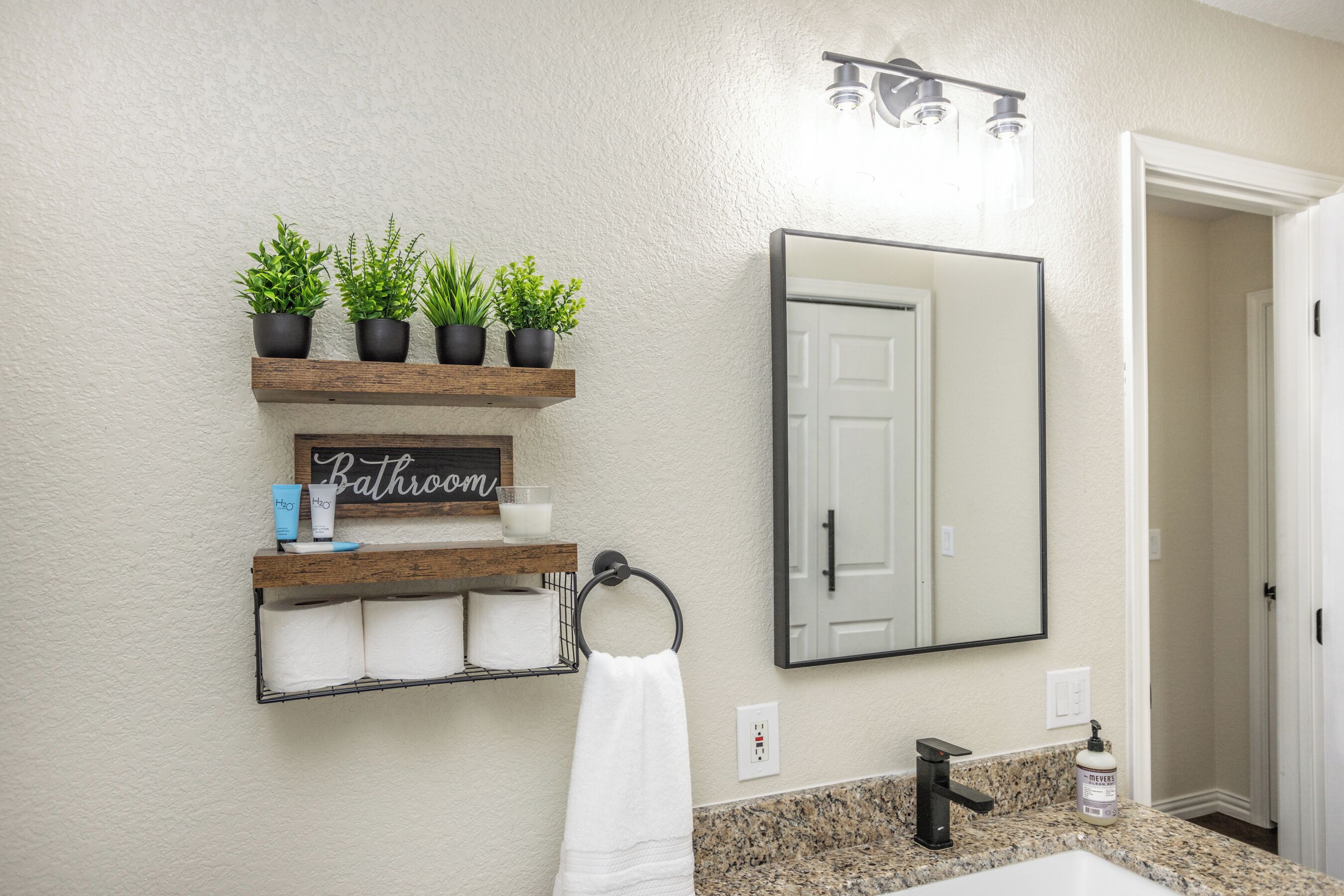 Un baño con un estante de madera con plantas en macetas, un perchero con una toalla blanca, un espejo y un lavabo con grifo.
