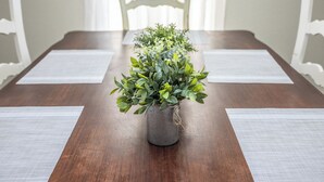 Une table à manger agrémentée d’une plante centrale placée dans un pot texturé, entourée de sets de table blancs et de chaises blanches aux dossiers décorés.