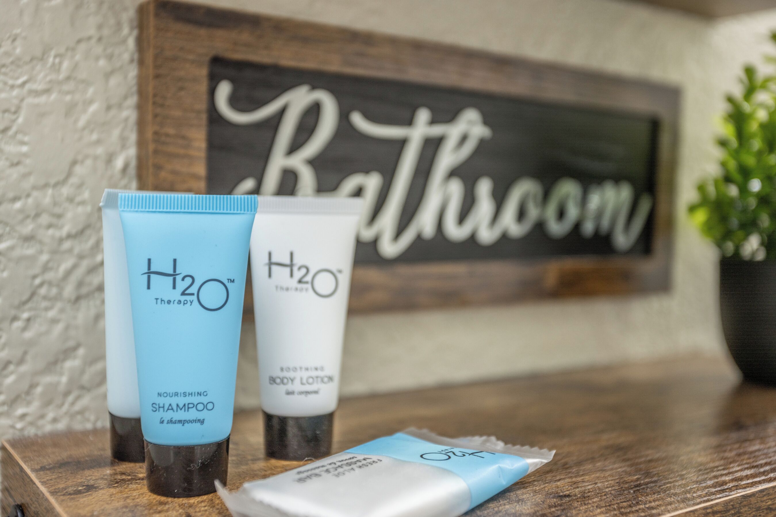 A bathroom vanity with a sign that reads "Bathroom" and three toiletry items: a blue H2O Therapy Nourishing Shampoo, a white H2O Therapy Body Lotion, and a blue H2O Therapy body wash.
