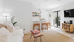 A modern living room with a white sofa, a coffee table, and a TV. There is a ceiling fan, a dining area with chairs, and a kitchen area with shelves. - Beachside Modern Wellness Villa with Spacious Patio, Walk to the Beach (Jupiter)