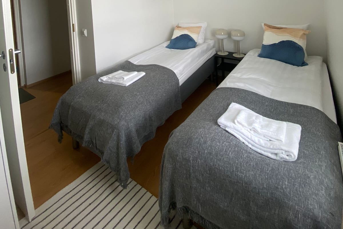 A room with two single beds, each with a grey blanket and a white pillow. There is a white pendant light hanging from the ceiling.