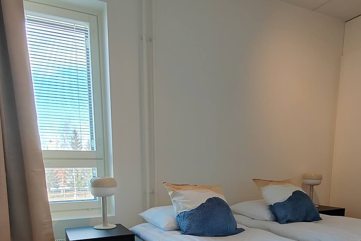 A bedroom with two beds, a nightstand, a lamp, and a window with blinds.