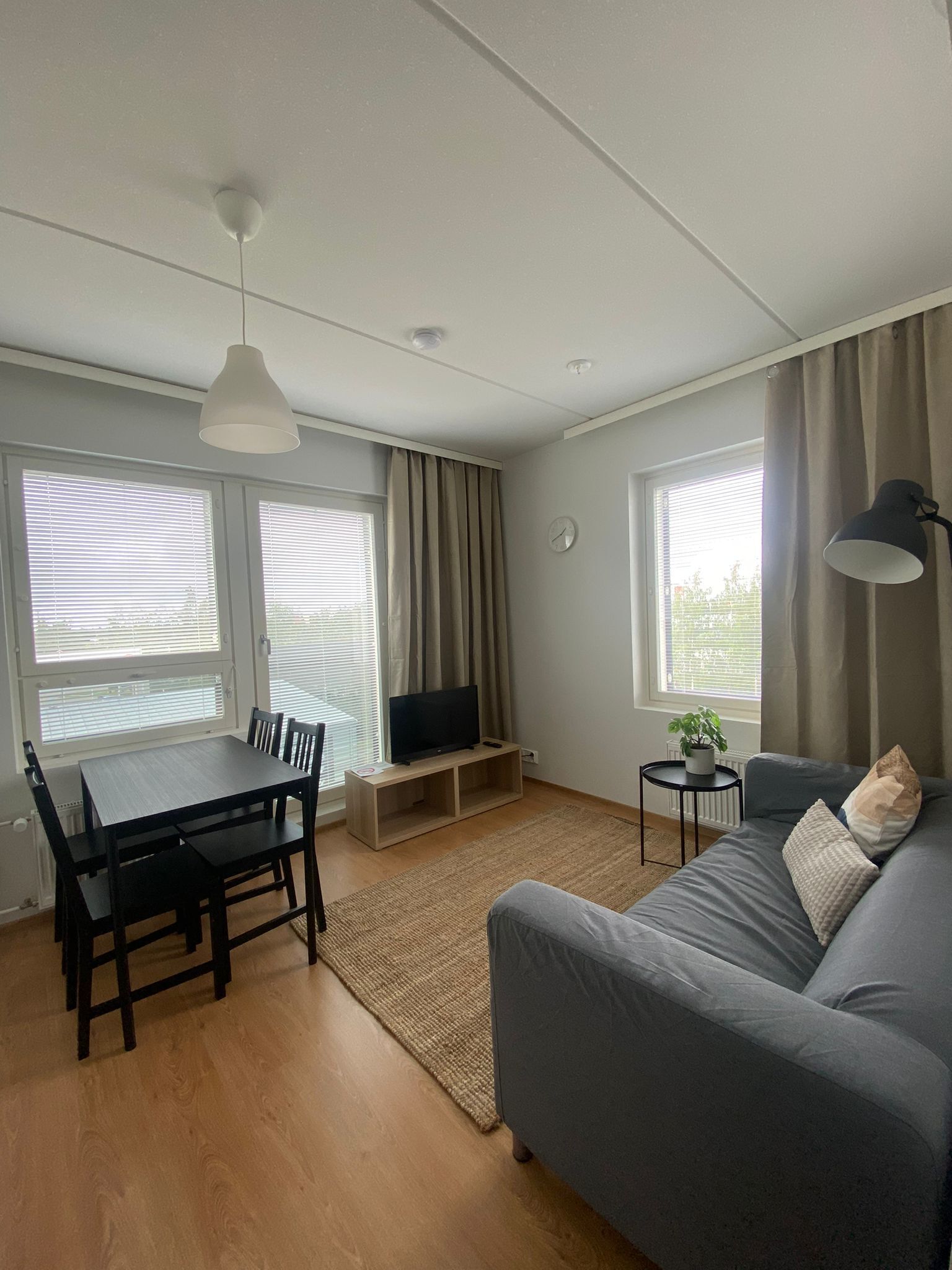 Una sala de estar con un sofá gris, un televisor sobre un mueble de madera, una mesa de comedor con sillas y una mesita auxiliar con una lámpara.