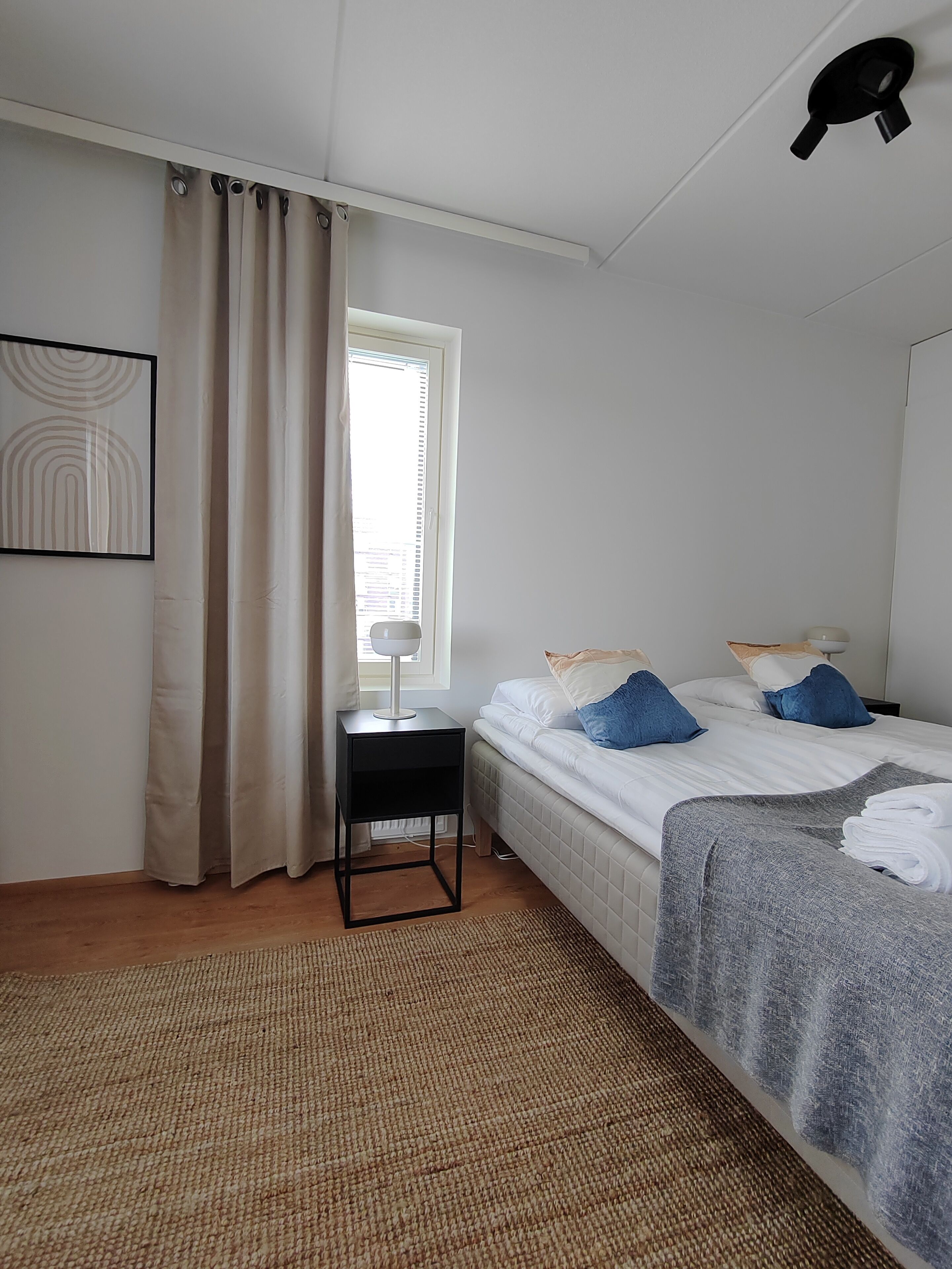 Une chambre à coucher comprenant un lit, une table de chevet, une lampe, une fenêtre avec des rideaux et un luminaire fixé au mur.