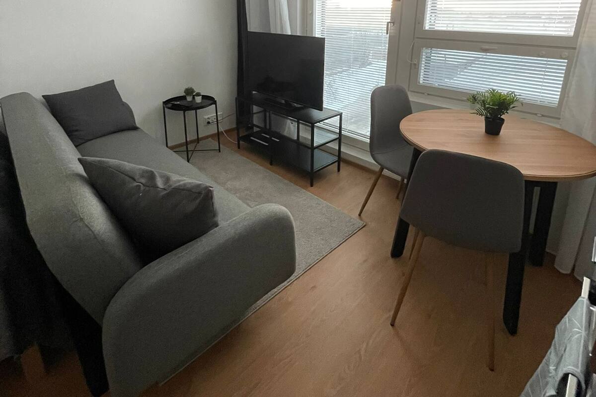 A modern living room with a grey sofa, a TV on a black stand, a dining table with chairs, and large windows with blinds.