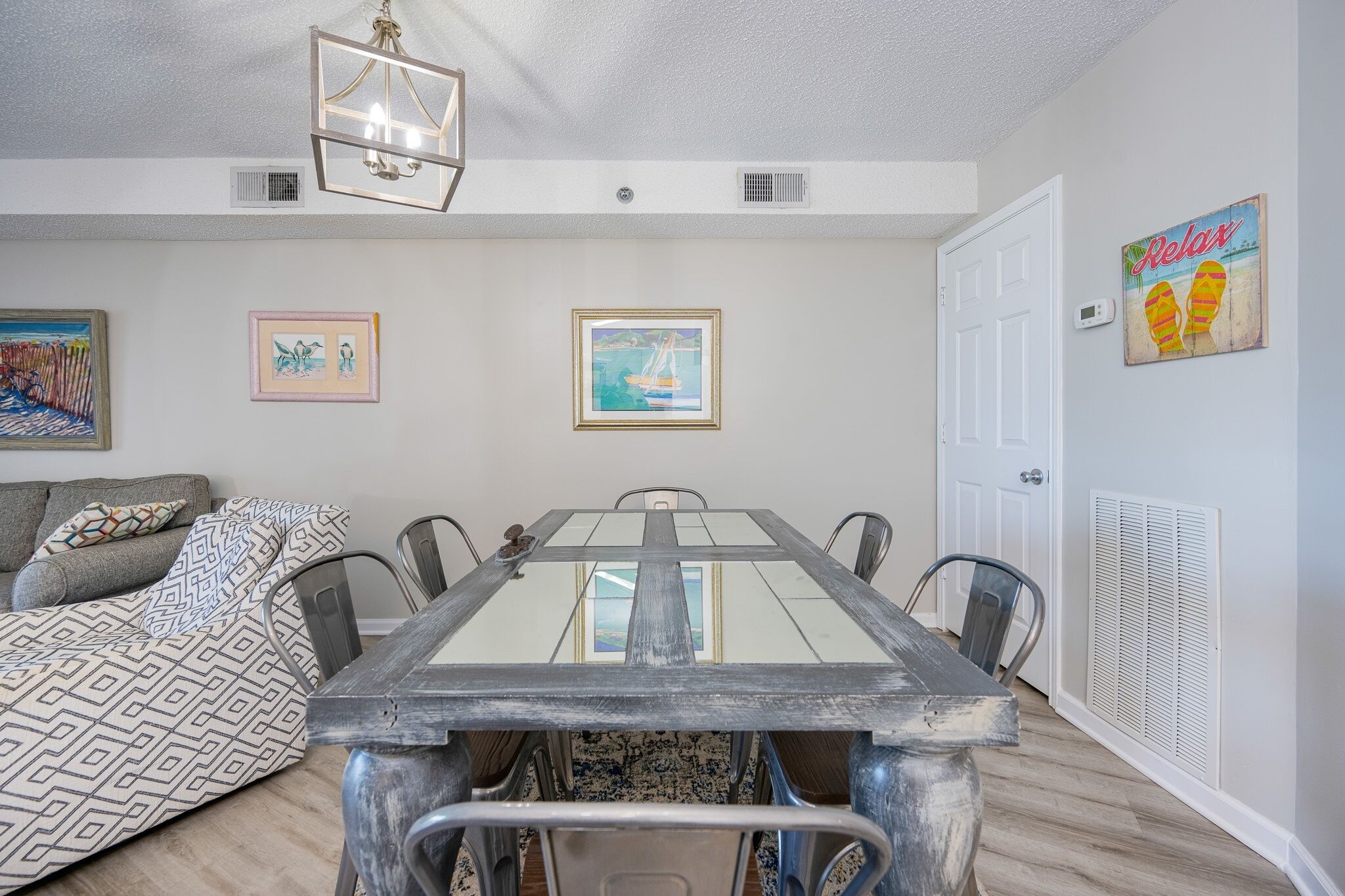 Un comedor con mesa de vidrio, sillas metálicas, un plaid estampado en el sofá y decoración en la pared.
