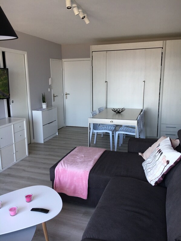 A modern, compact living space with a sofa, a small table with candles, a dining area with a table and chairs, and a kitchenette with cabinets and a TV.
