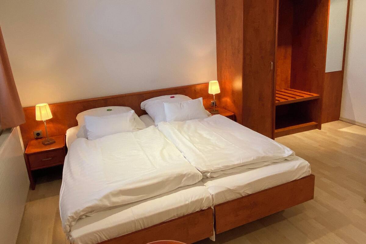 Ein ordentlich bezogenes Doppelbett mit weißen Laken, zwei Nachttischlampen und ein Holzschrank mit Spiegel.
