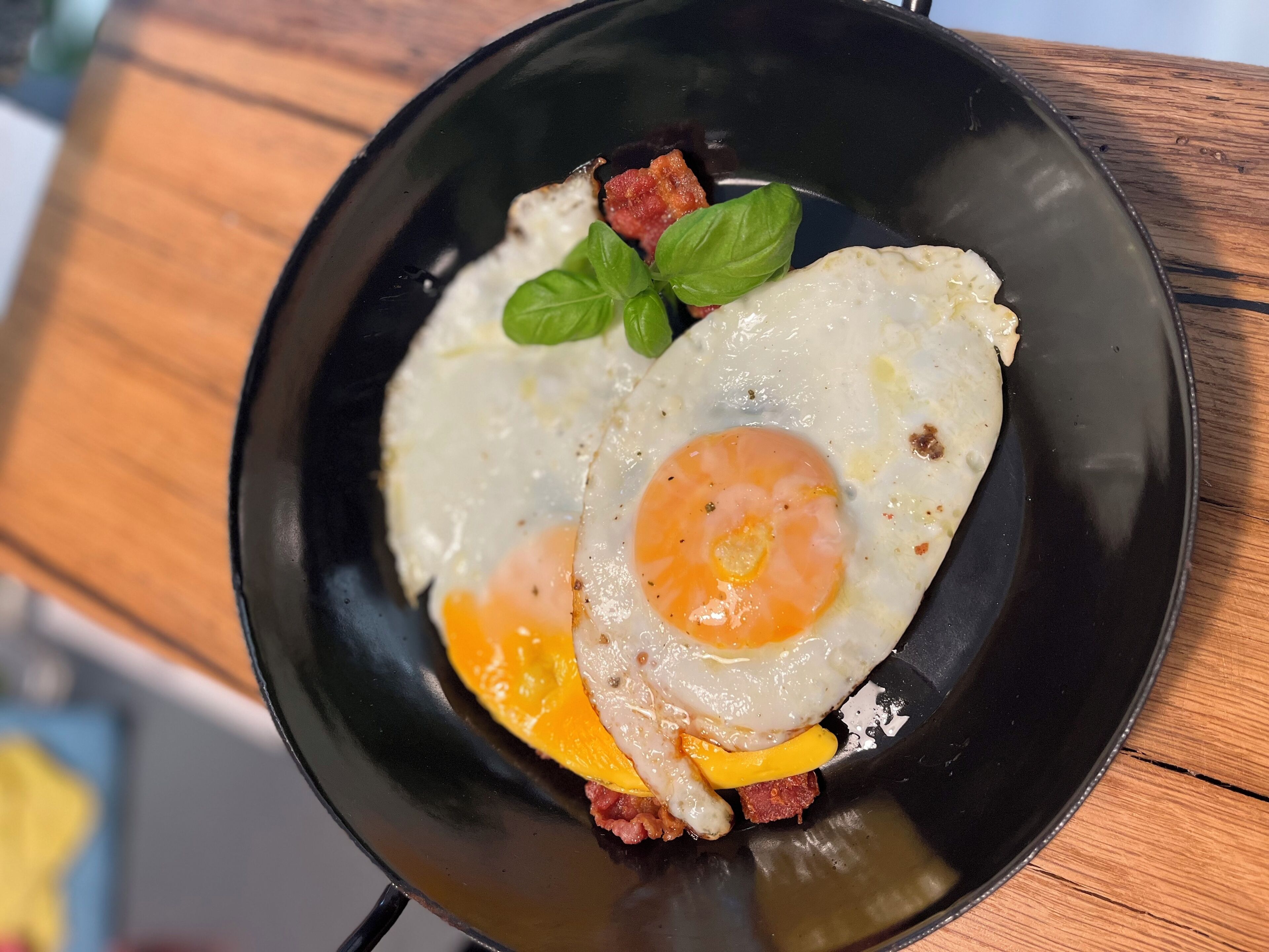 A black skillet with two sunny-side-up eggs, a slice of yellow cheese, and a piece of red meat on a wooden table.