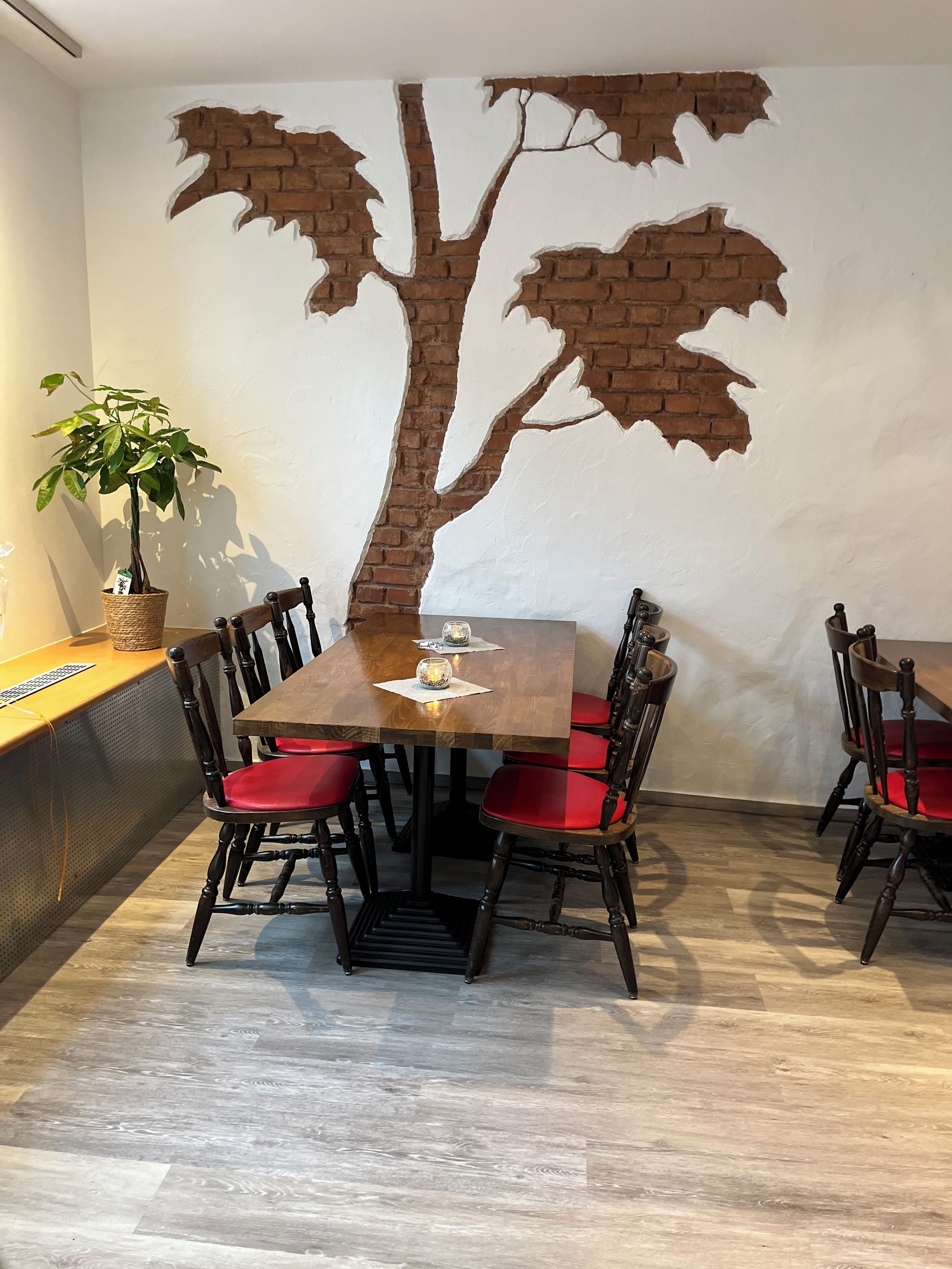 Un coin repas avec une table en bois et des chaises à revêtement rouge, une plante en pot et une fresque murale représentant un arbre.